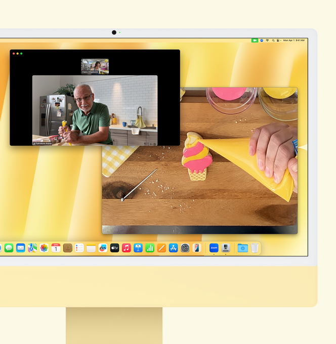 iMac amarelo. Demonstração do recurso Visualização da Mesa. A tela mostra uma pessoa em uma confeitaria fazendo uma chamada de vídeo com outra pessoa. Uma das telas mostra as pessoas da chamada de vídeo. A outra tela mostra as mãos da pessoa que está fazendo a cobertura de um cookie.