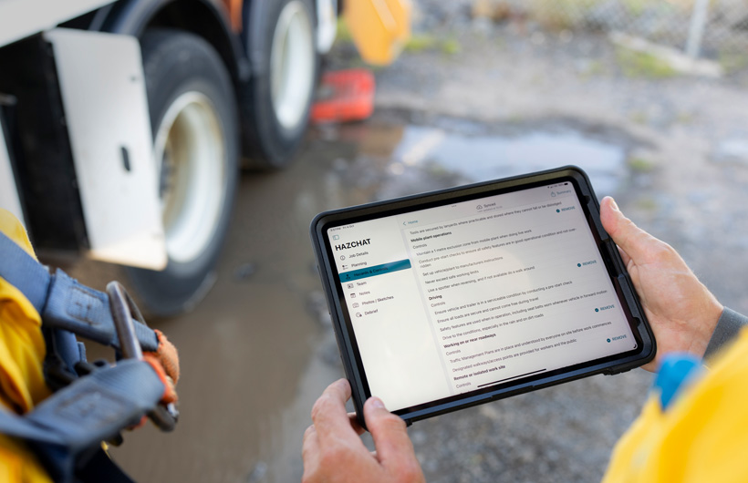Hands hold an iPad with the HazChat app open which displays Job Details, Planning, Hazards and Controls, while a person stands at a work site by a mud puddle